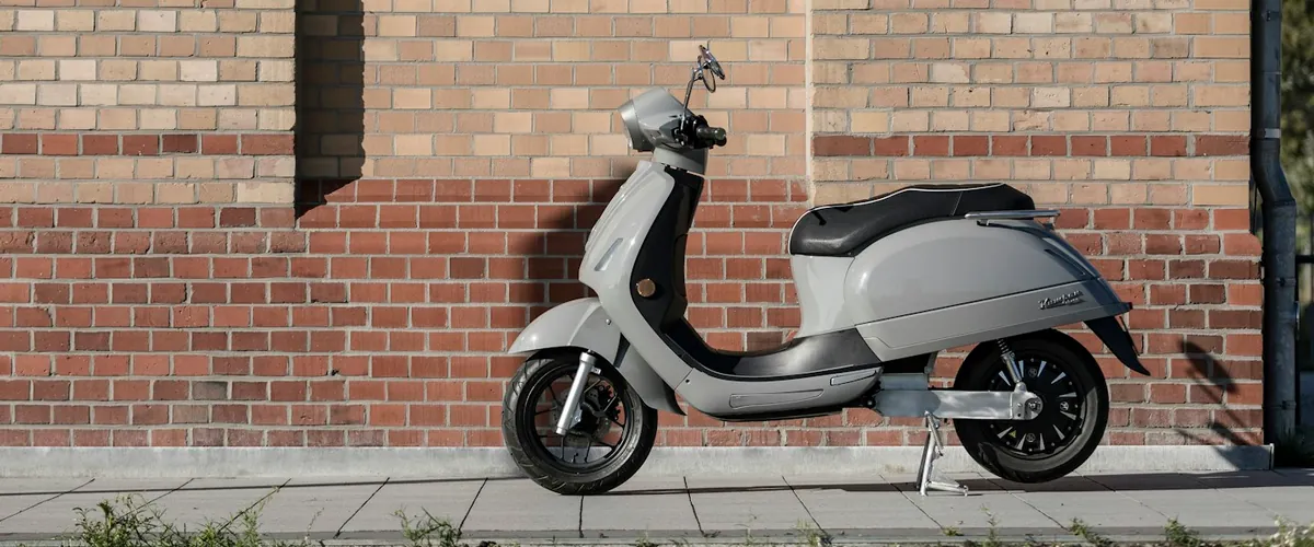 Scooter eléctrico gris estacionado junto a una fachada de ladrillo; fotografía de contexto